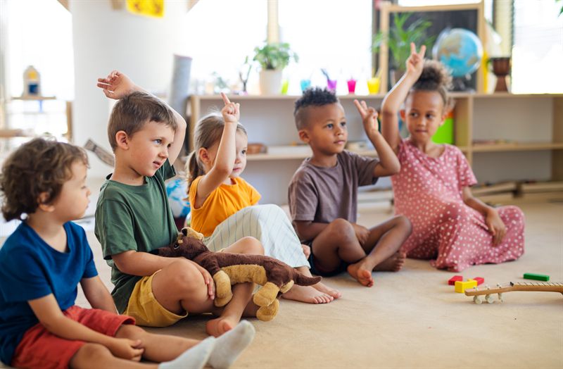 Nursery children sitting together on the floor with their hands raised during a group activity